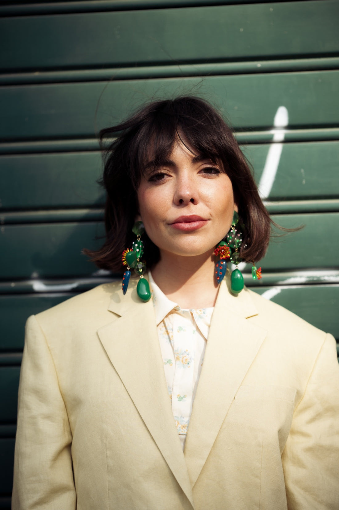 Woman in a tailored city outfit wearing statement earrings in front of olive shutters