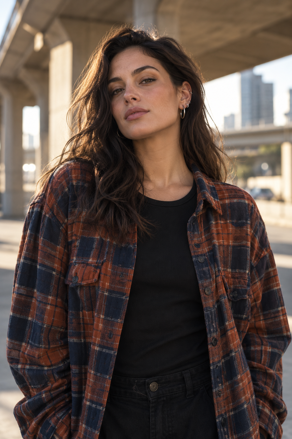 Adult model in a rust and navy plaid overshirt with a silver ear stack beneath a city underpass