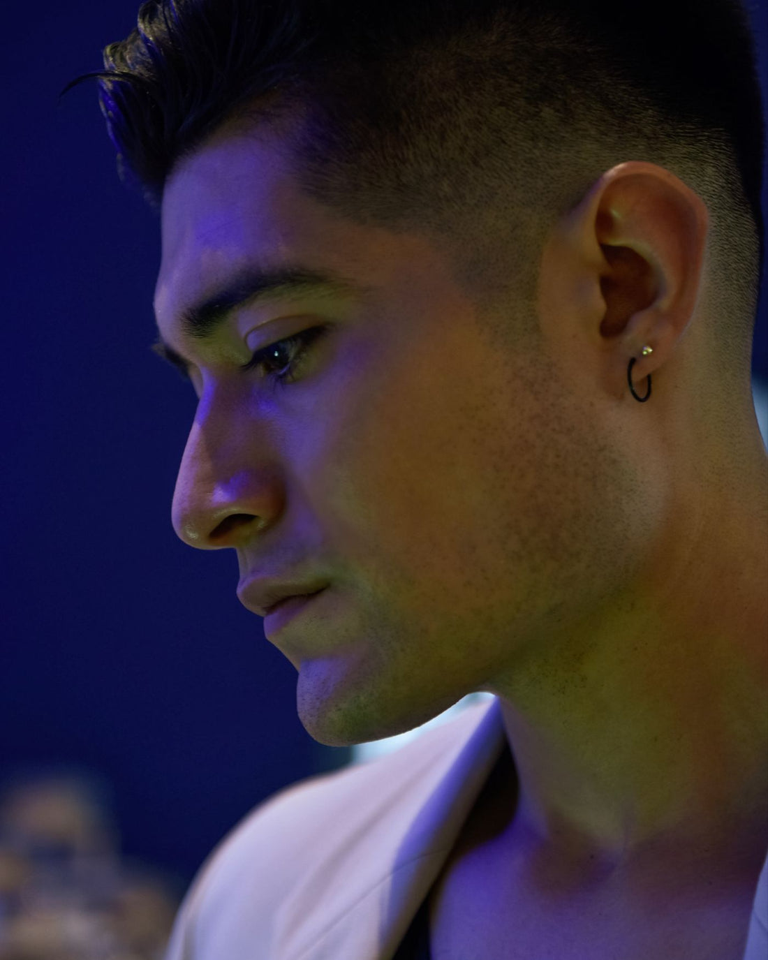 Blue low-light portrait of a model wearing a small ear hoop with dark streetwear styling