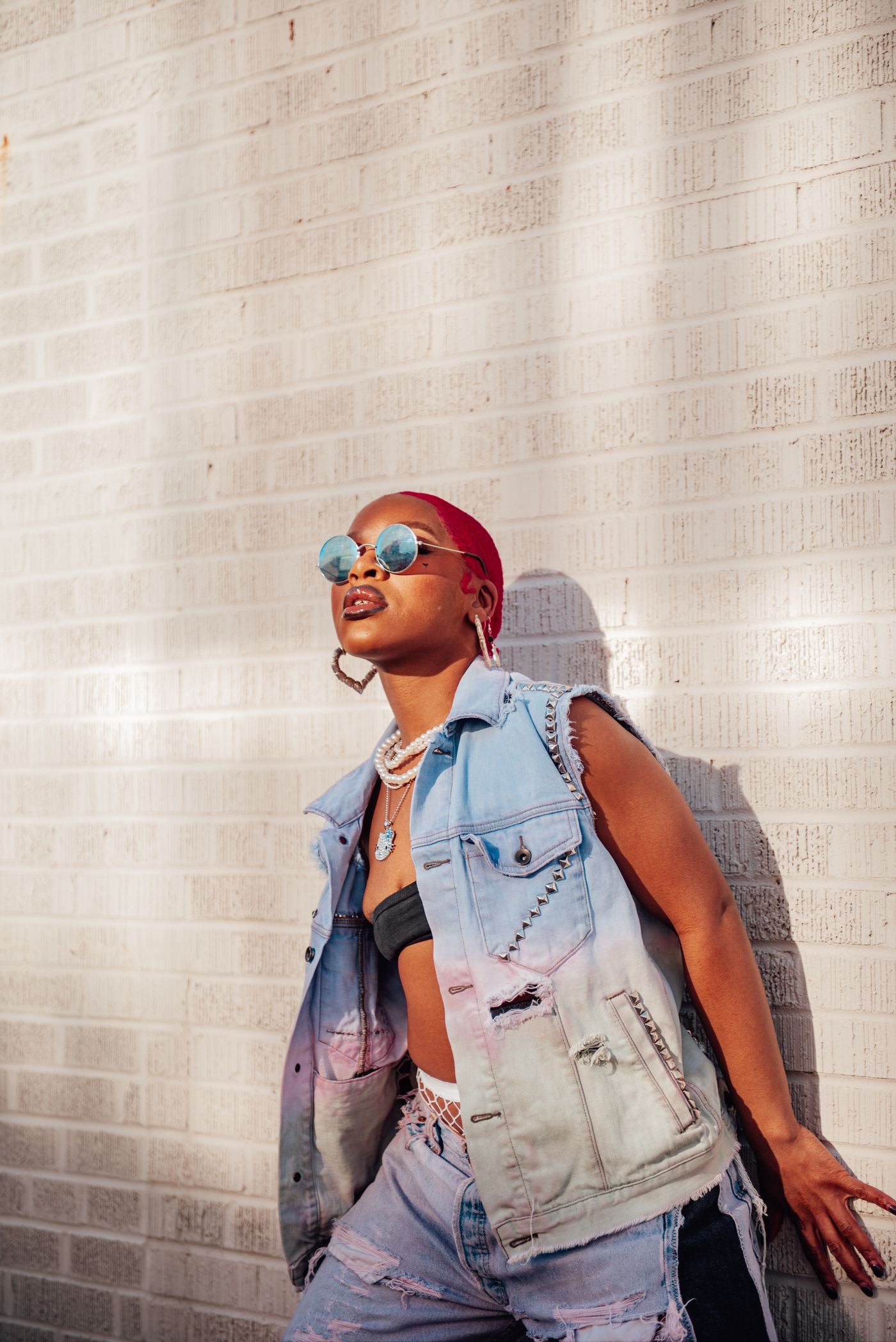 Model with pink hair wearing a denim vest and sunglasses against a brick wall