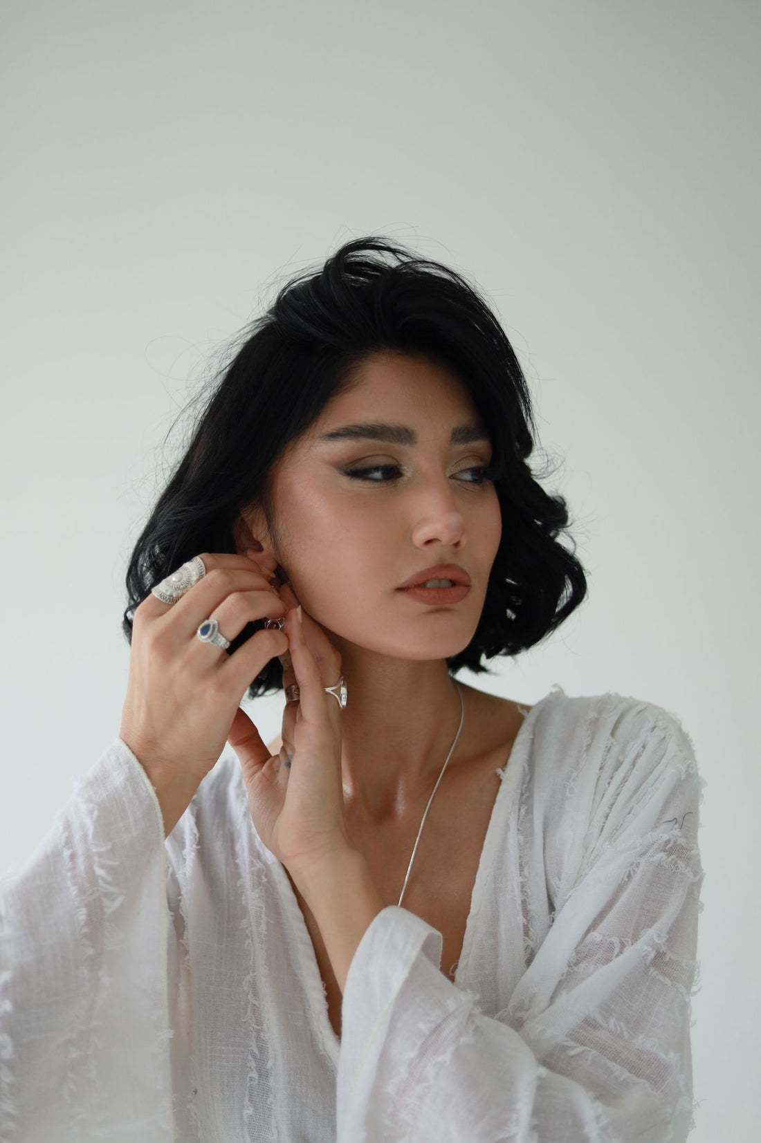 Woman in soft white layers adjusting earrings and wearing silver rings for an evening jewelry look